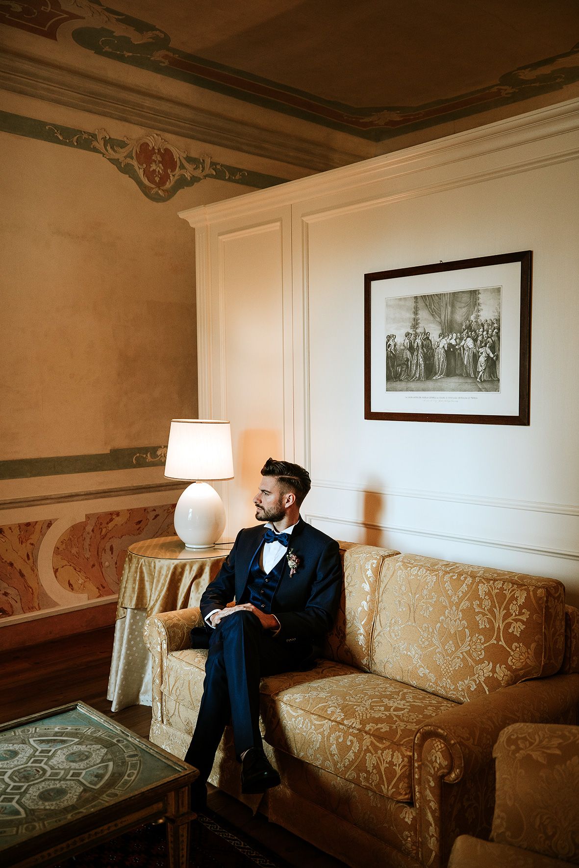 Person in formal attire sits on vintage gold sofa in elegant room with ornate ceiling and framed artwork.