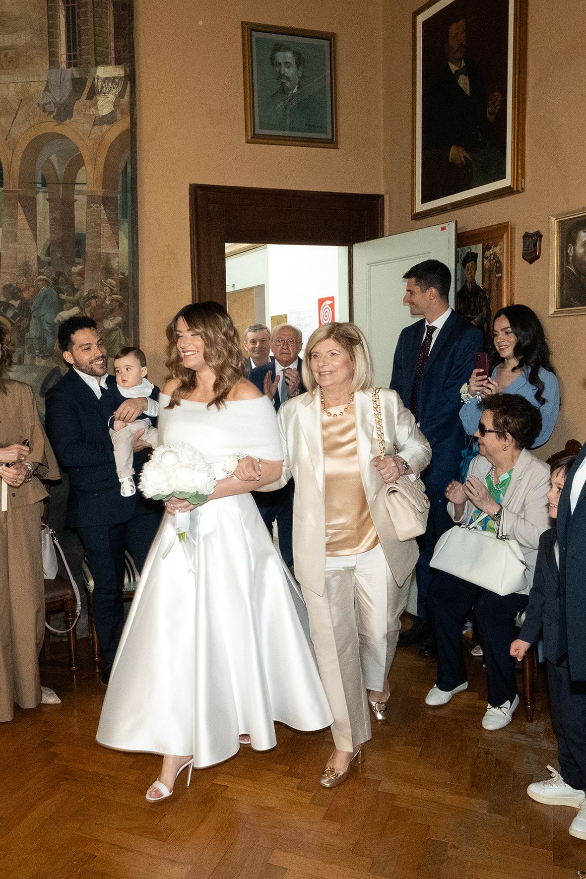 Wedding party gathers in an elegant room with hardwood floors as a bride in a white tea-length dress walks with guests.