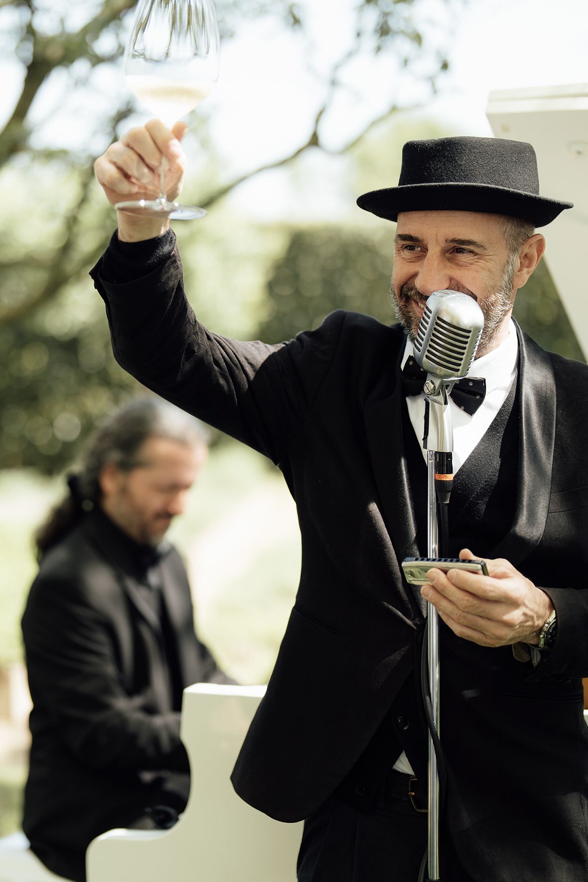 A performer in a black suit and hat raises a glass while singing into a vintage microphone at an outdoor event.