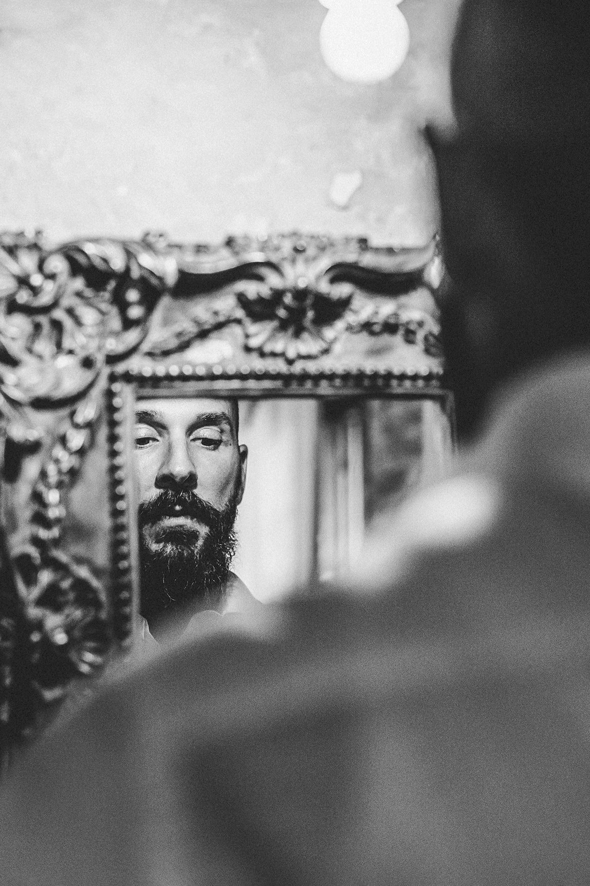 Black and white reflection in ornate mirror shows bearded person against decorative frame with intricate details.