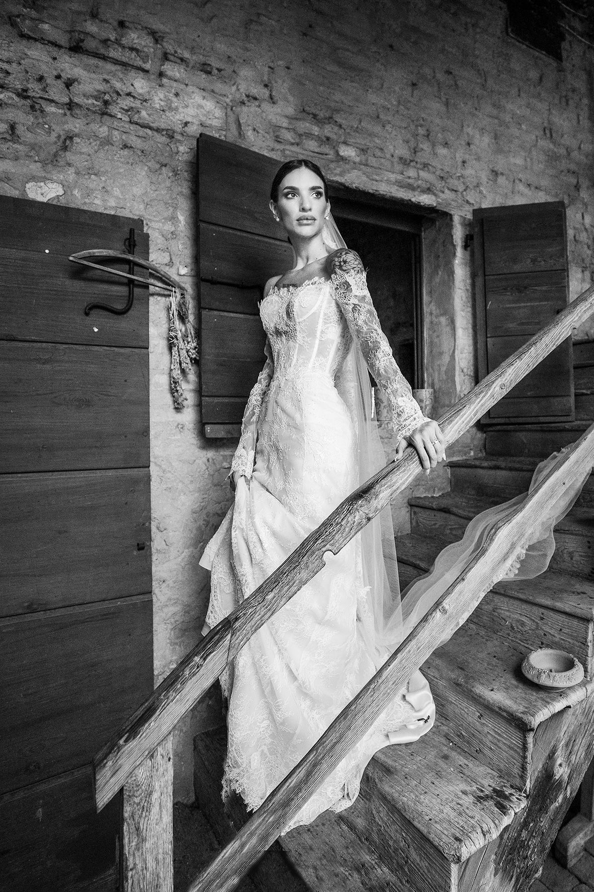 A vintage black and white photo showing a person in an elegant lace wedding dress standing on rustic wooden stairs.