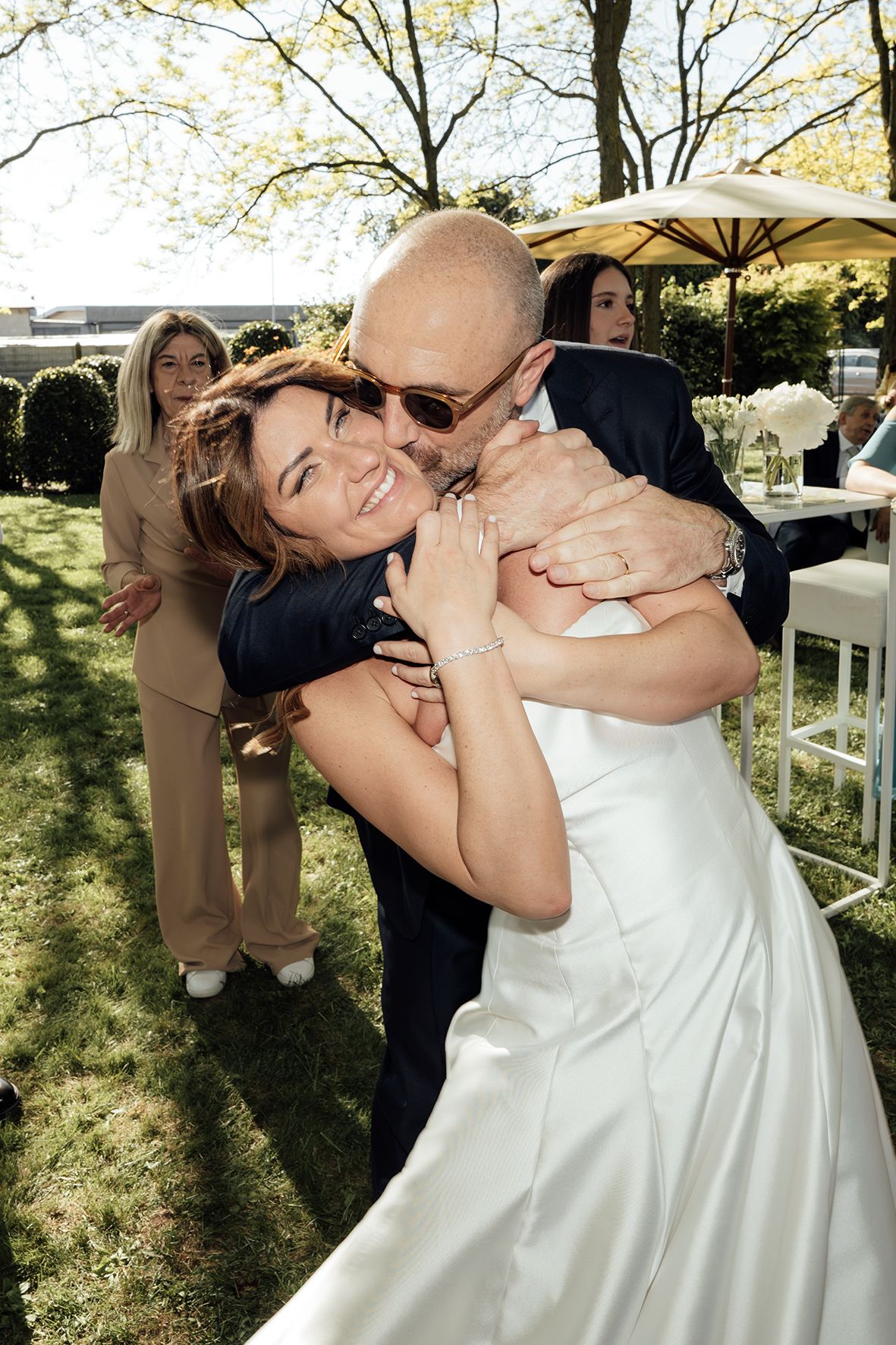 Two people embrace joyfully at an outdoor wedding reception with white chairs and umbrellas visible in the garden setting.