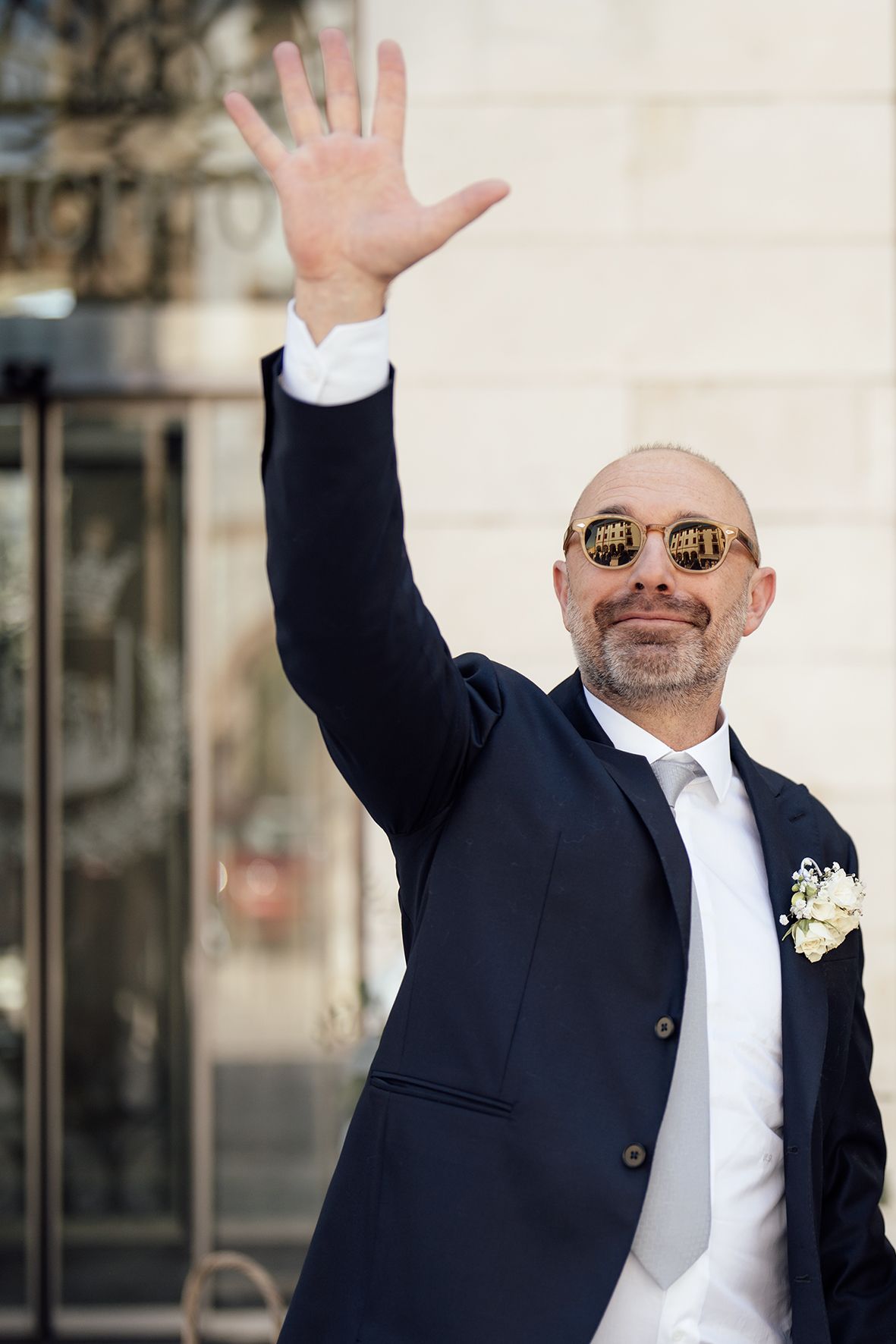 A well-dressed man in a navy suit and sunglasses waves to the crowd while wearing a boutonniere.