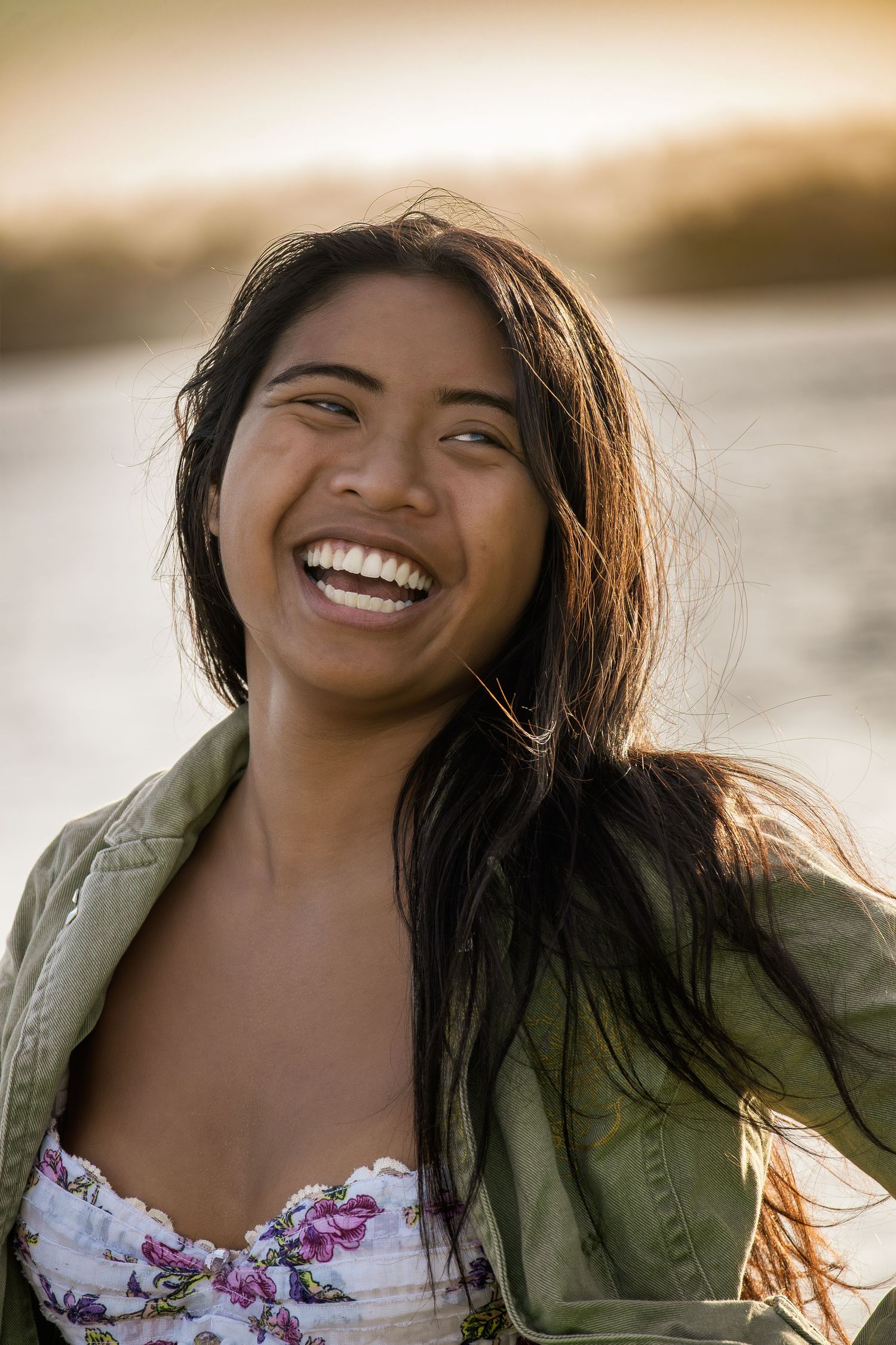 A person with long dark hair wearing a floral top and green jacket laughs joyfully in warm sunlight.