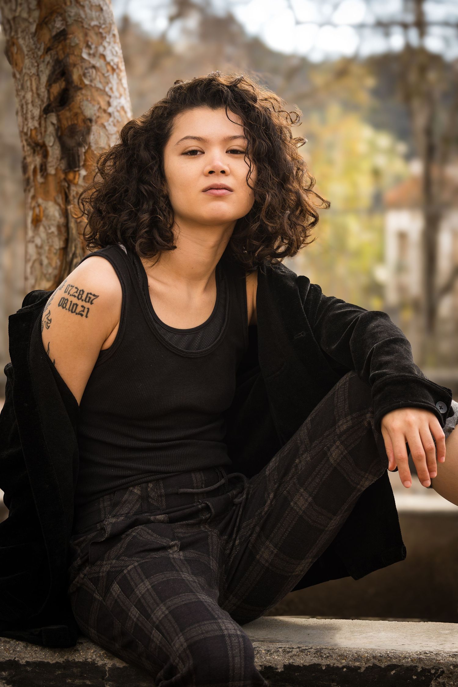 A person in black clothing with curly hair reclines on a stone surface in an outdoor autumn setting.