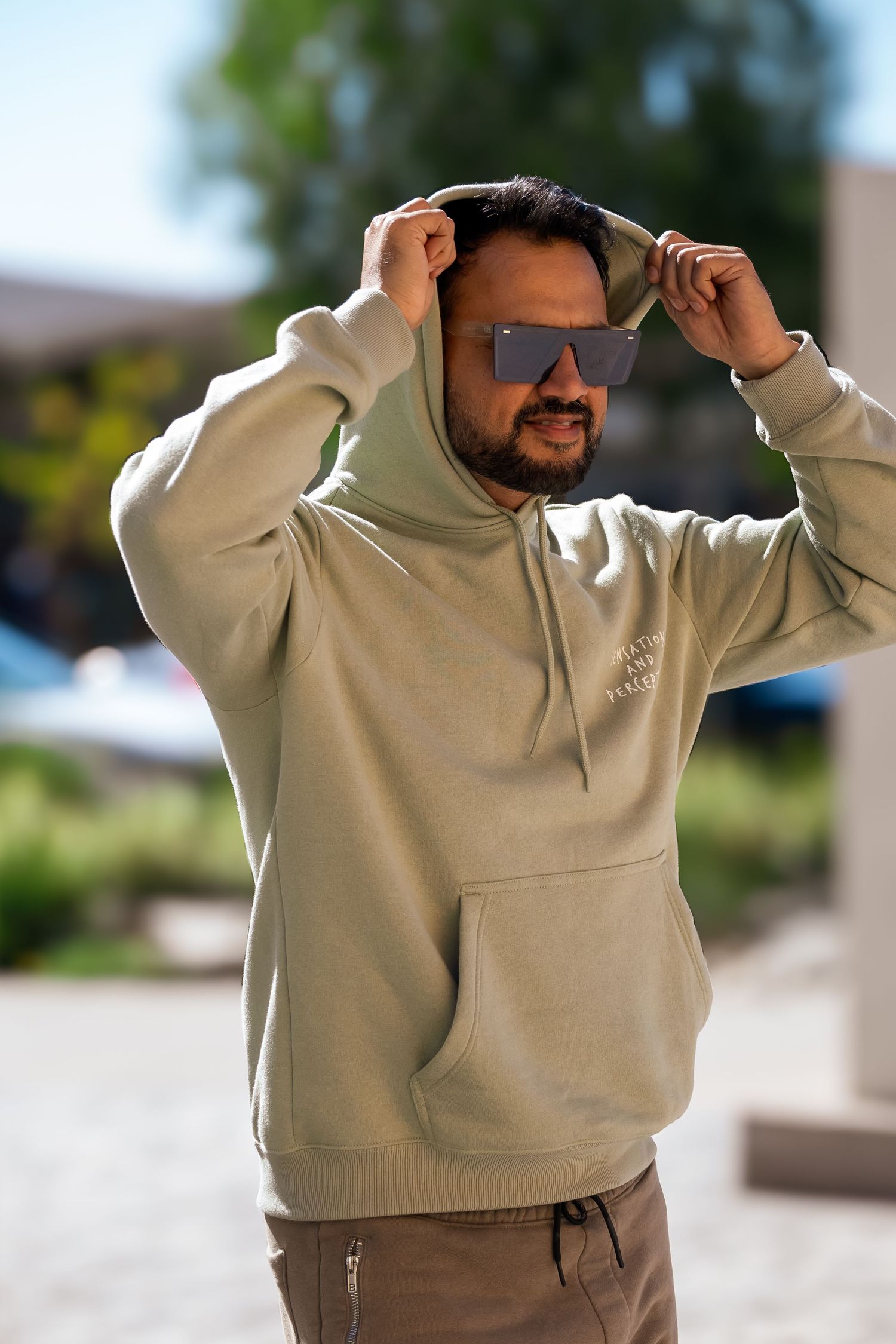 Person in beige hoodie striking playful poses outdoors on sunny day.