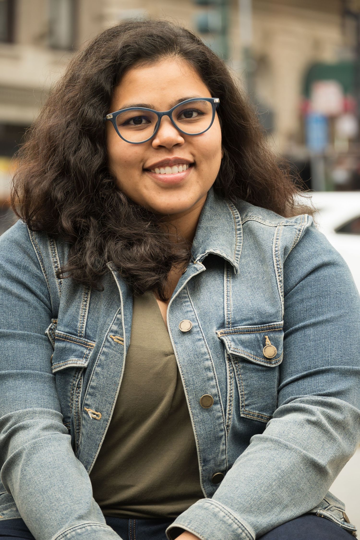 A person wearing glasses and a denim jacket smiles warmly while sitting outdoors on a sunny day.