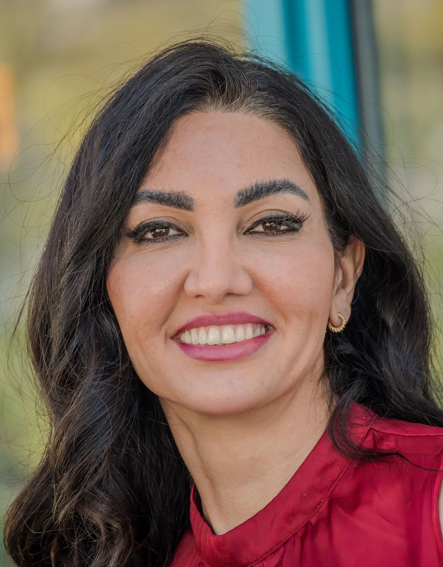 A smiling woman with long dark hair wearing a red top poses for a professional headshot against a blurred background.