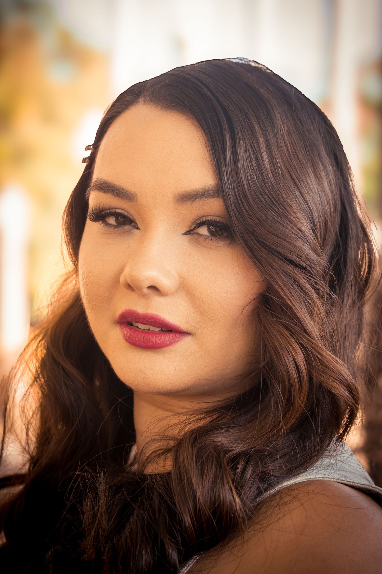 Portrait of a young woman with dark wavy hair and red lipstick, posing outdoors in warm sunlight.