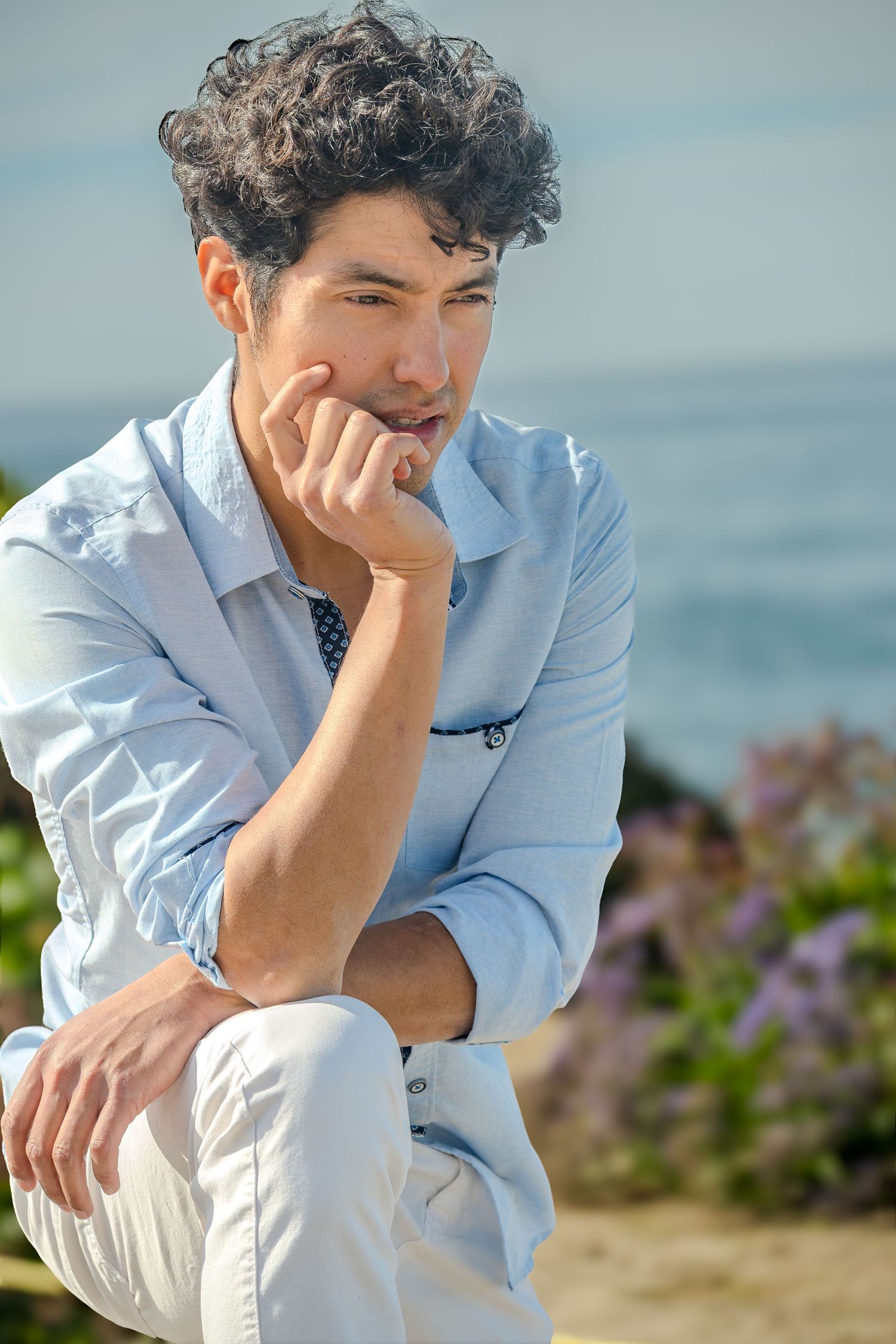A person in a light blue shirt sits thoughtfully by the ocean with purple flowers in the background.