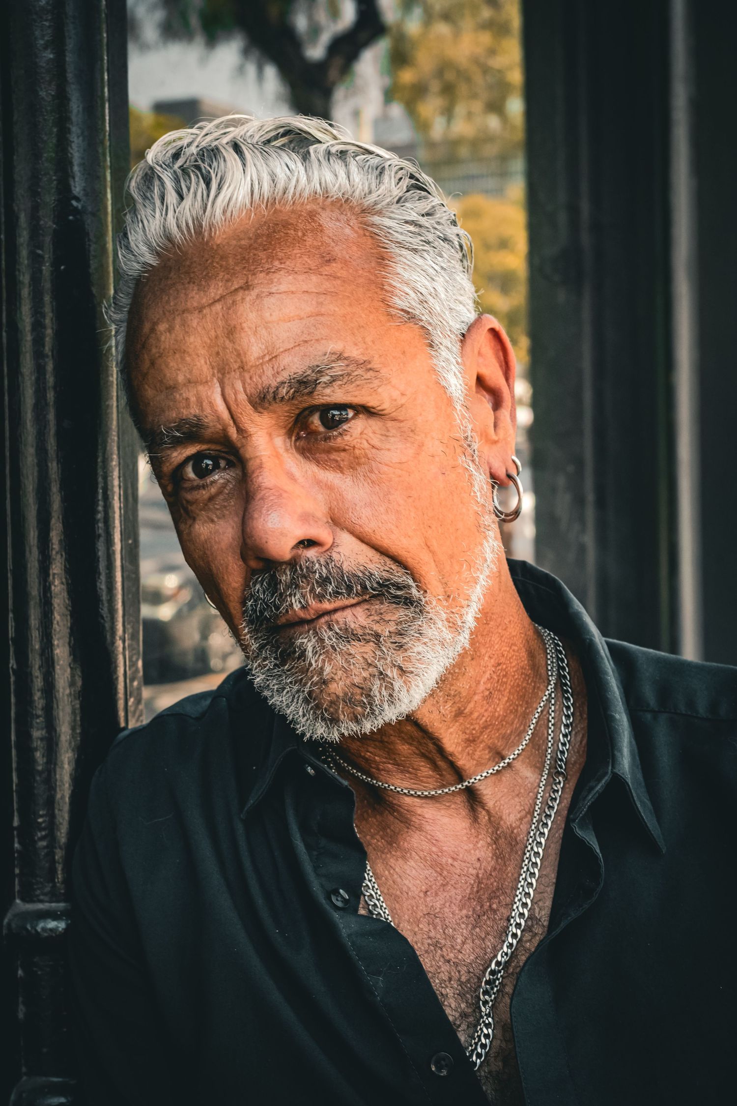 Portrait of a mature person with silver hair and jewelry wearing a dark shirt against an architectural background.