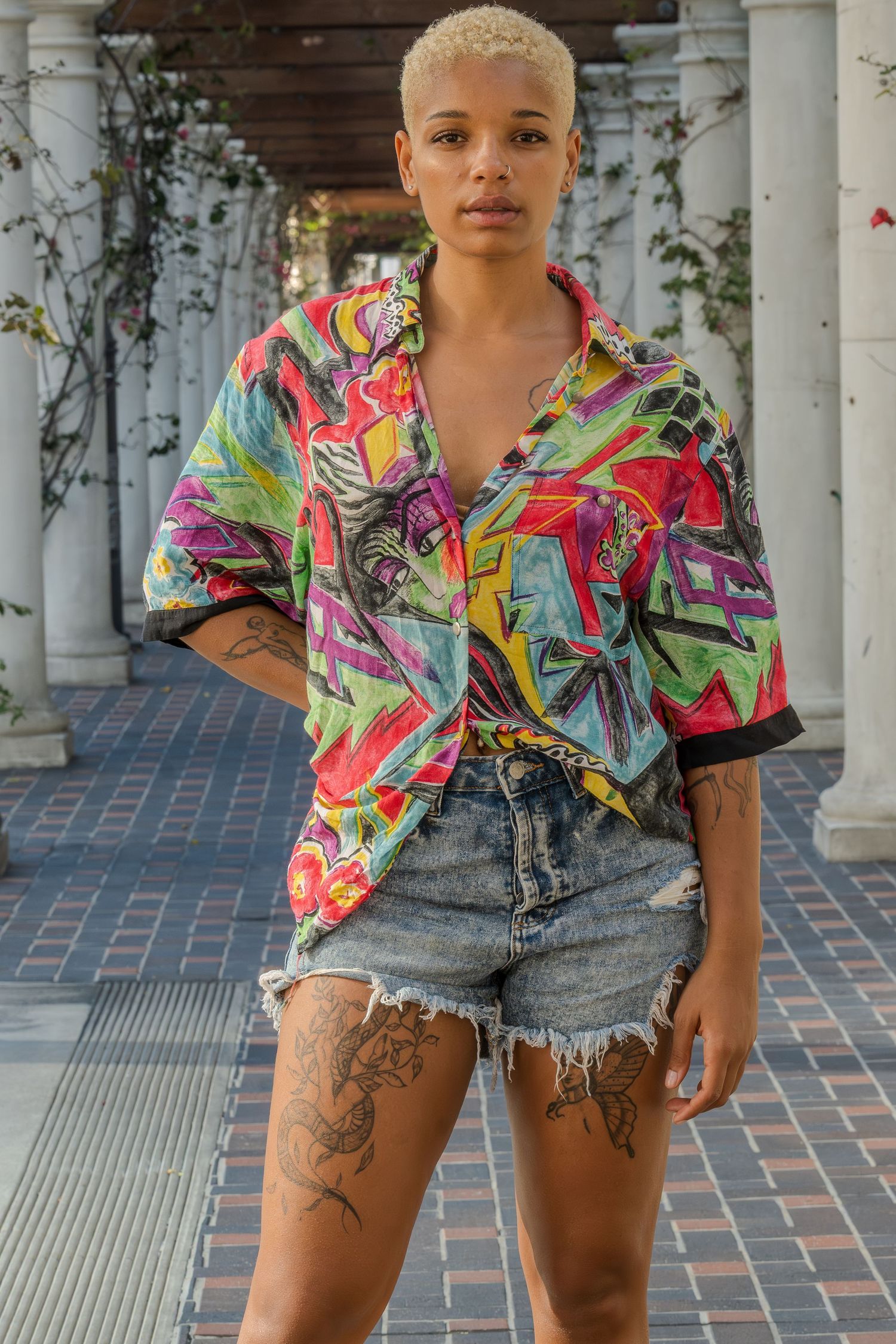 A person wearing a colorful abstract print shirt and distressed denim shorts poses in four similar shots against white columns.