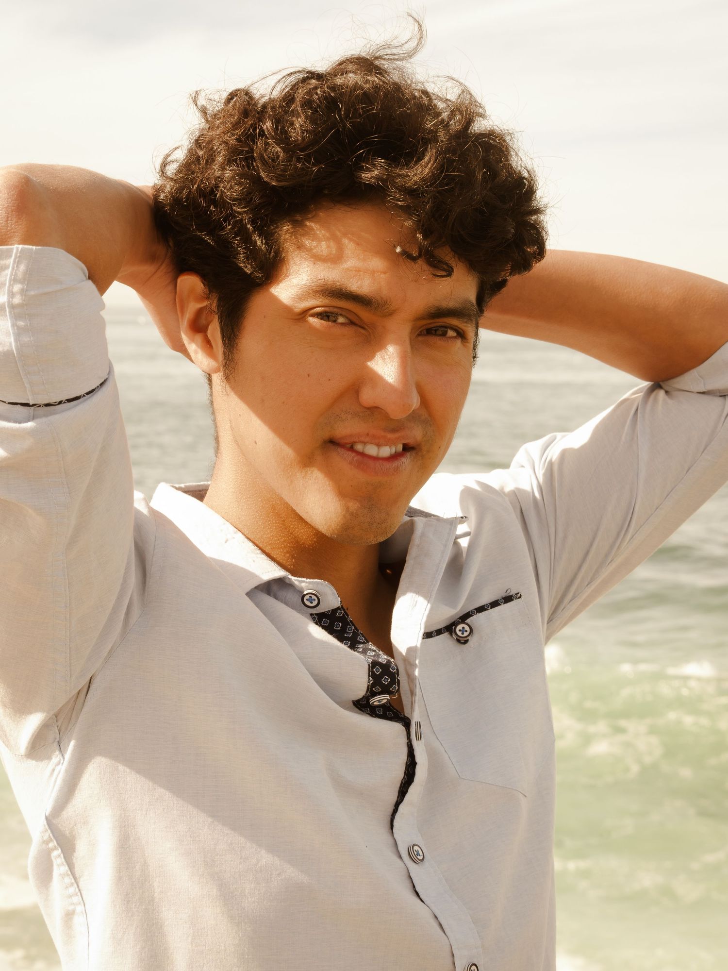 Person in white shirt posing with hands in hair against bright sunlit background.