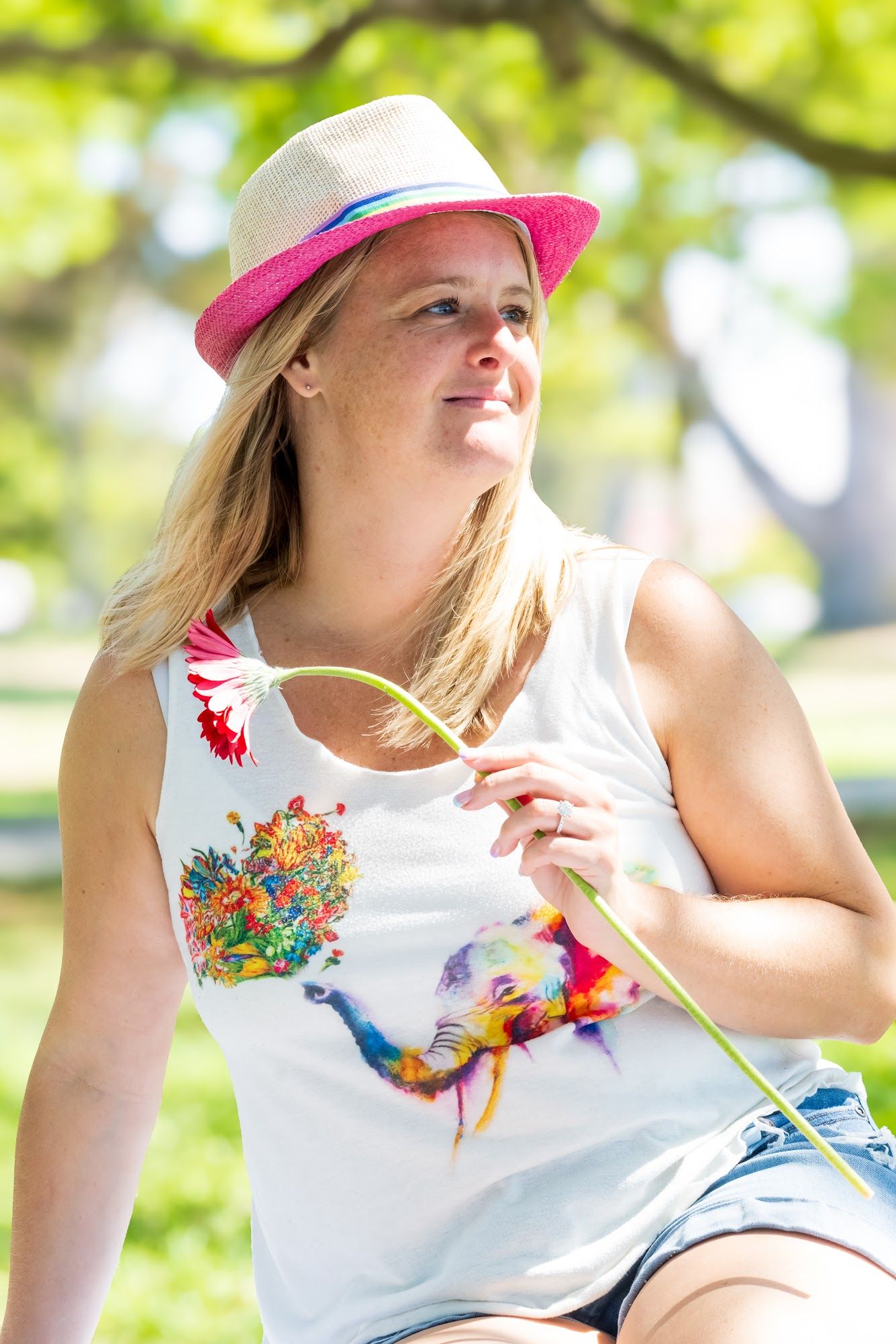 Person wearing a colorful bird print tank top and pink-brimmed hat poses in a sunny outdoor setting.