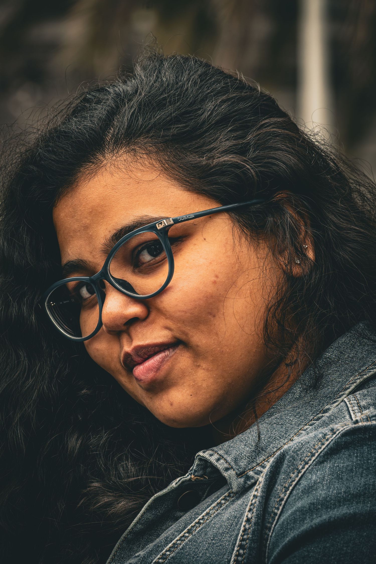 Close-up portrait of a person wearing black-rimmed glasses and a denim jacket, with a soft smile, against a blurred background.