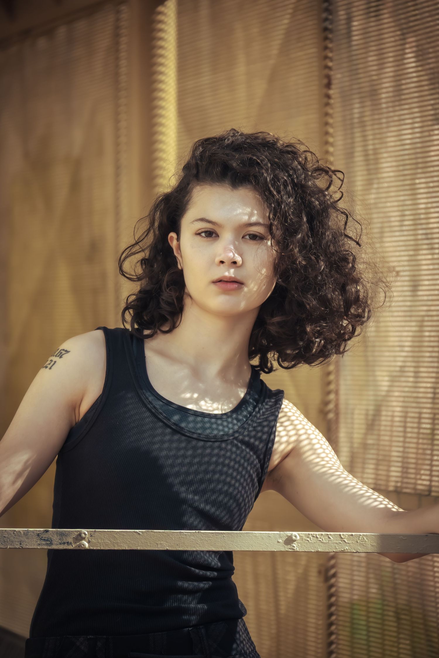 A person in a black sleeveless top poses dramatically against venetian blinds with sunlight streaming through.