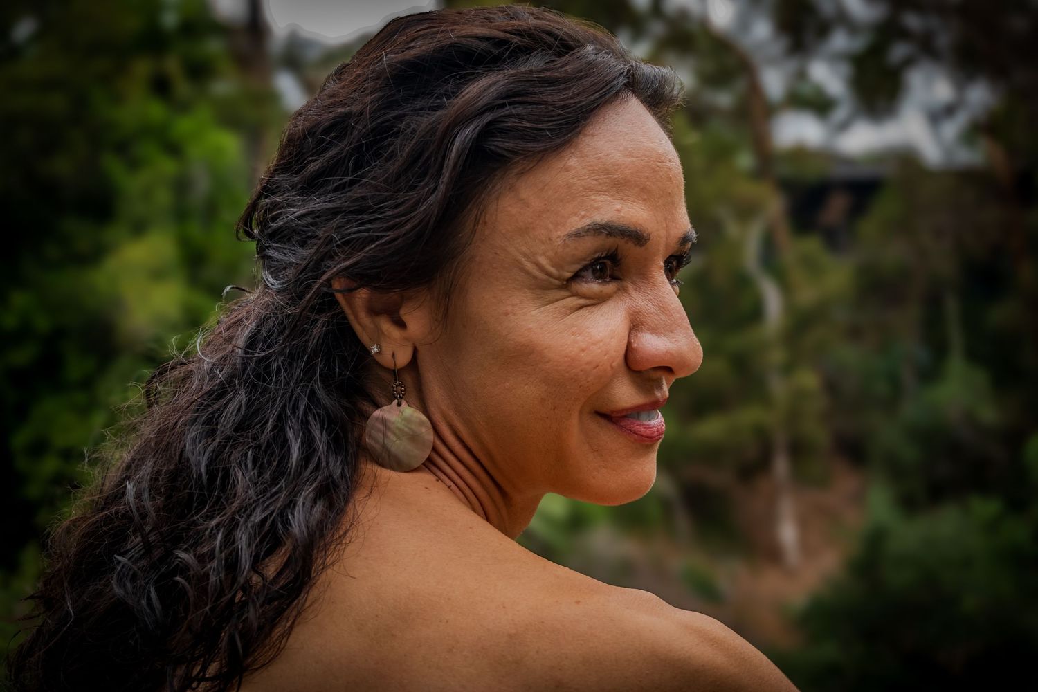 A smiling woman with dark wavy hair and large round earrings poses outdoors with lush green trees in the background.