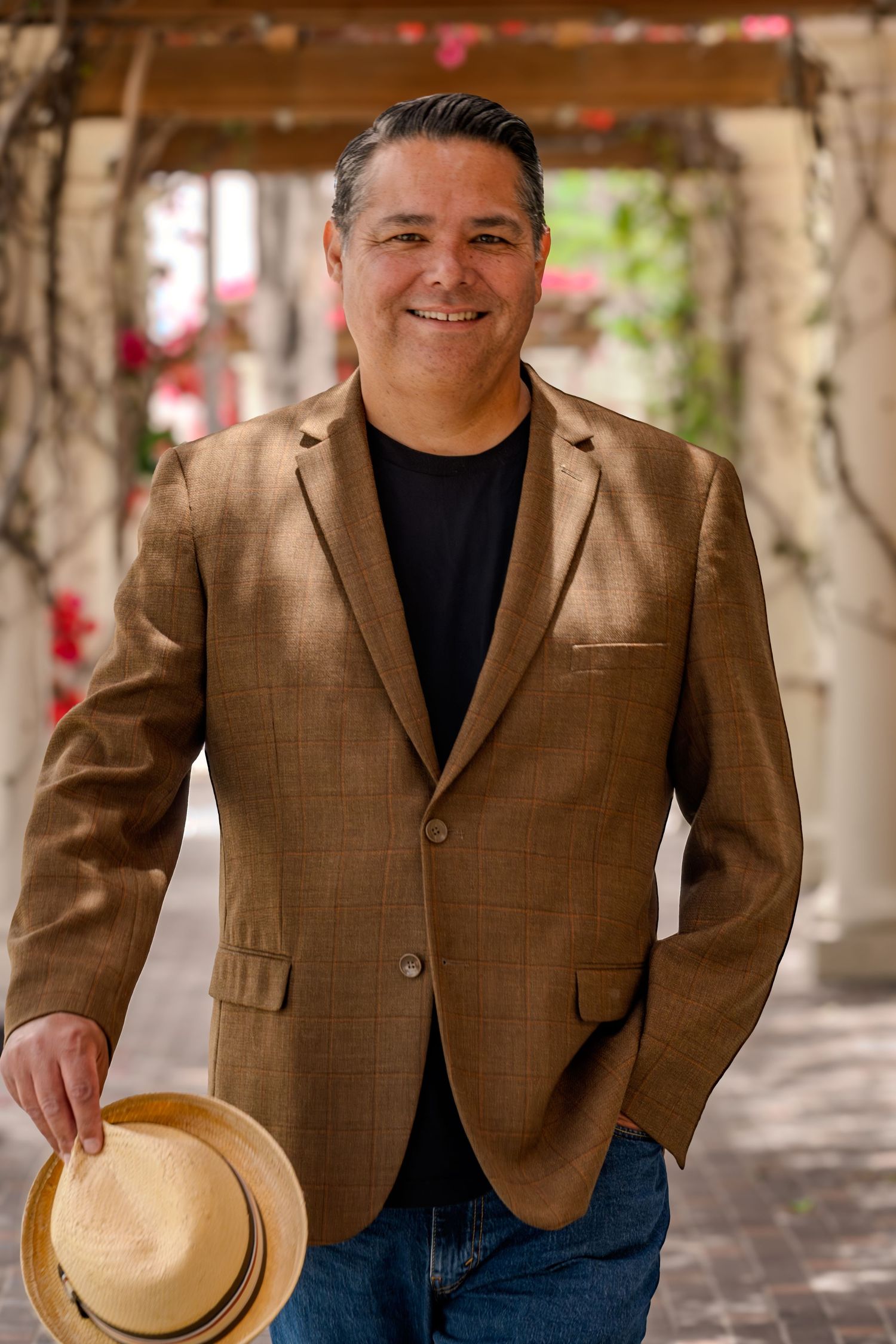 A person wearing a brown blazer and black turtleneck poses outdoors with a straw hat against a blurred background.