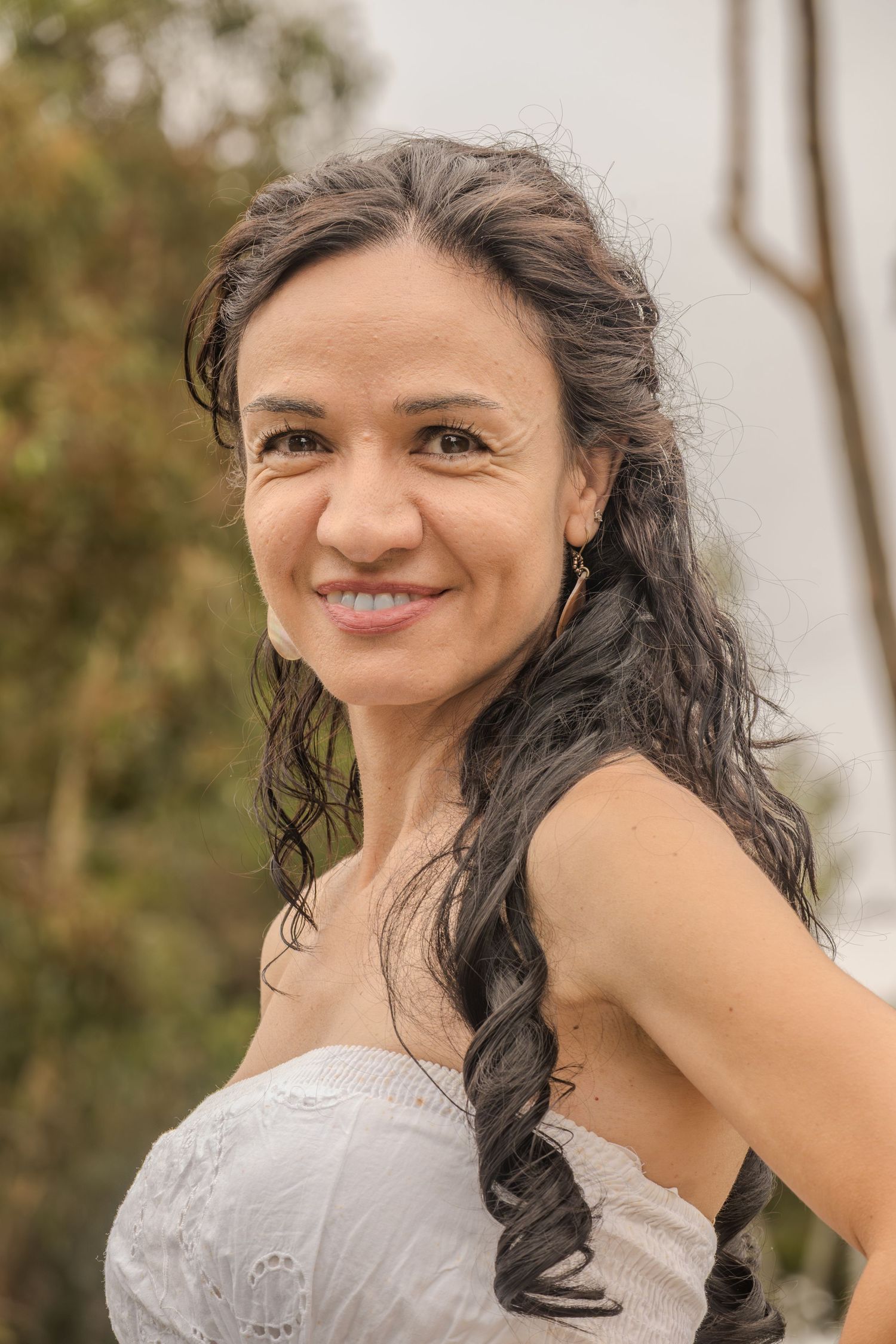 Smiling middle-aged woman with long dark hair wearing a white strapless top outdoors.