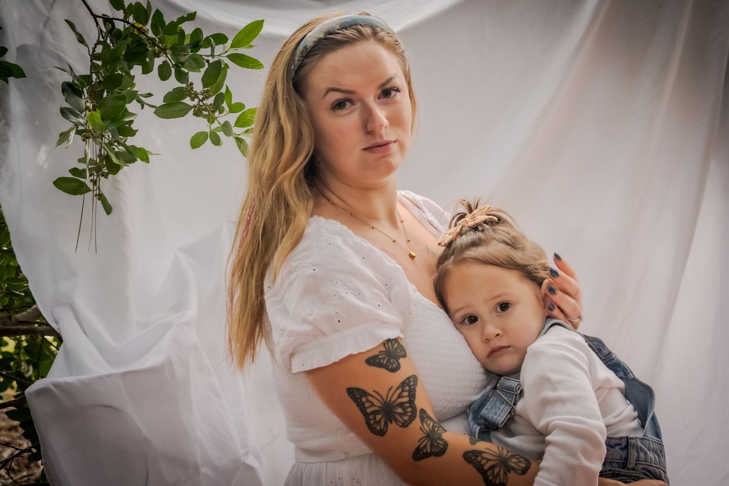 Mother and child in white clothing pose together for intimate portrait session with natural greenery backdrop.