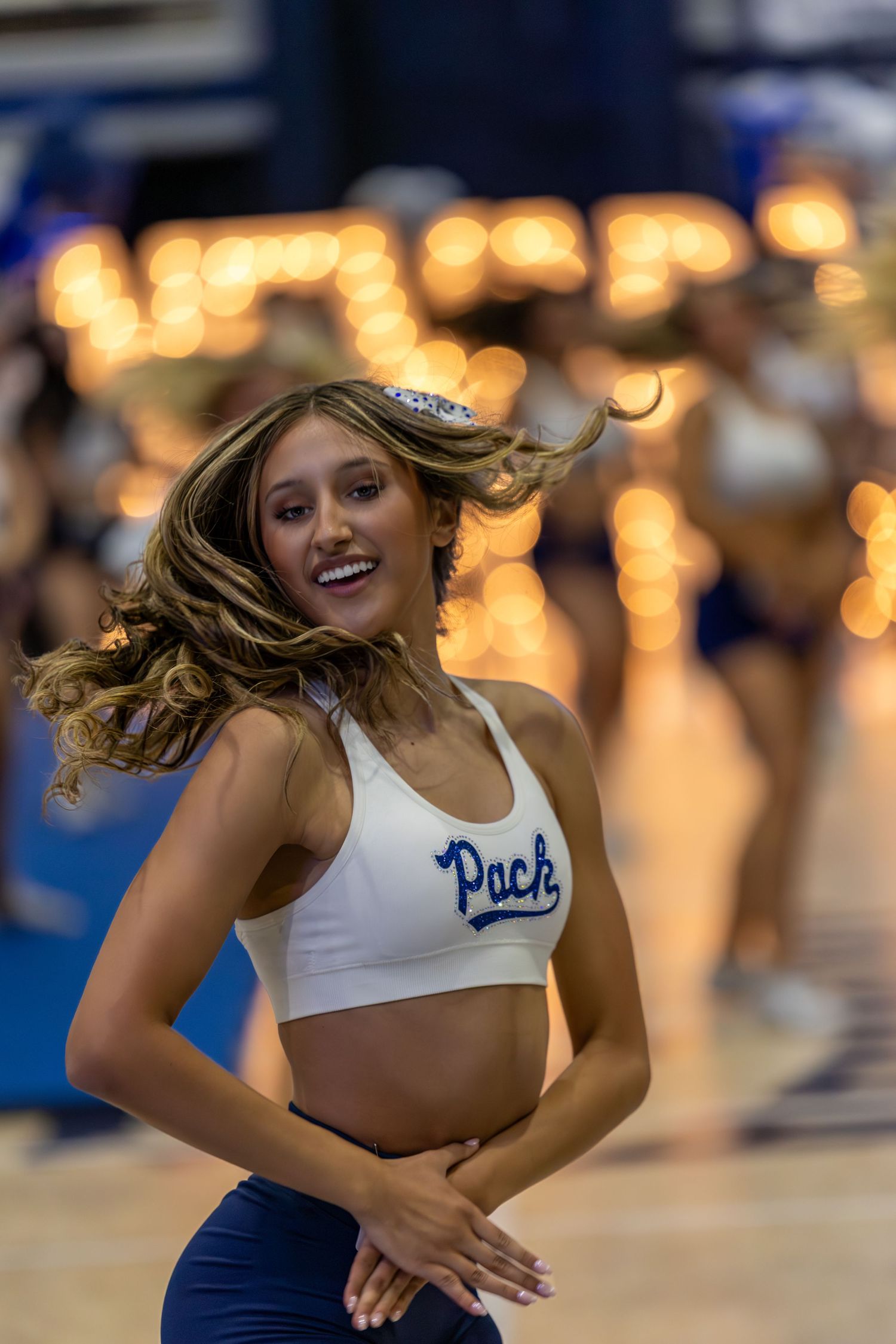 A cheerleader in a white Pack crop top smiles while dancing at a university sports event with bokeh lights in the background.