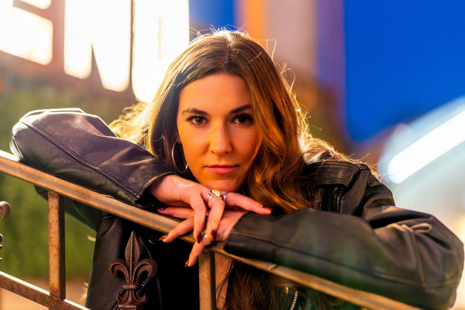 Person in a leather jacket poses against a railing with dramatic lighting and blue background.