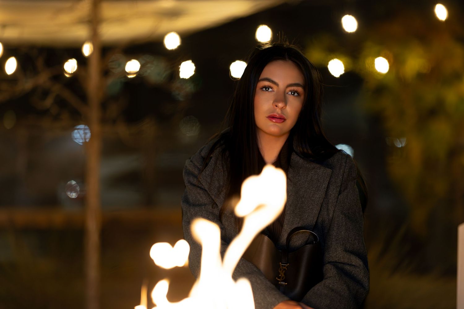 Young woman in gray coat standing near open fire at night with string lights glowing in background.