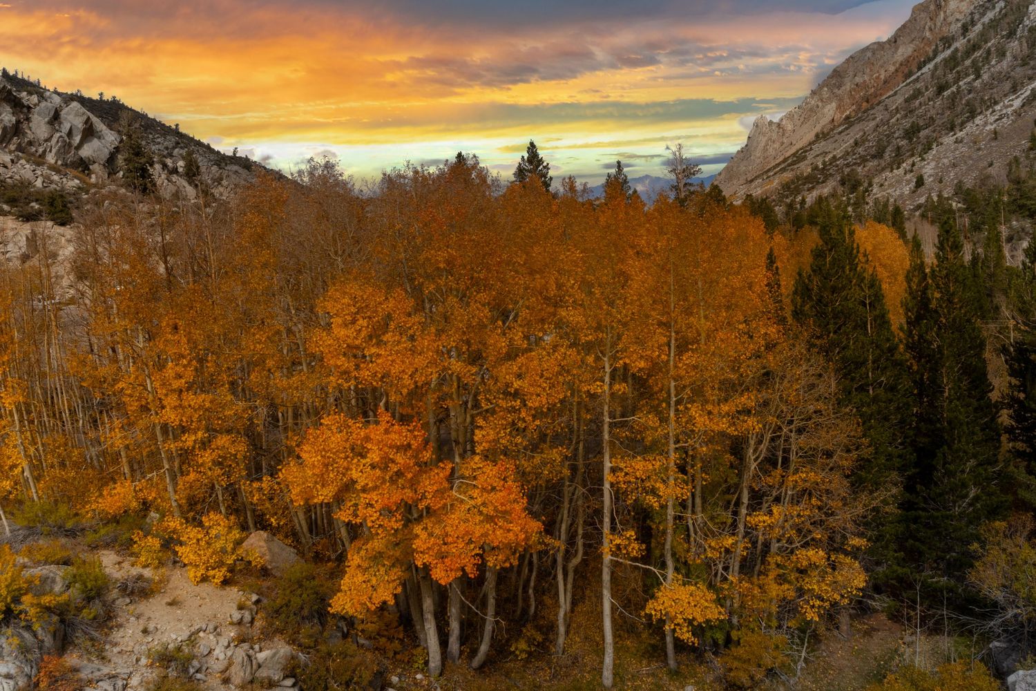 Scenic mountain landscape with golden aspen trees and dramatic sunset clouds illuminating the rugged peaks.