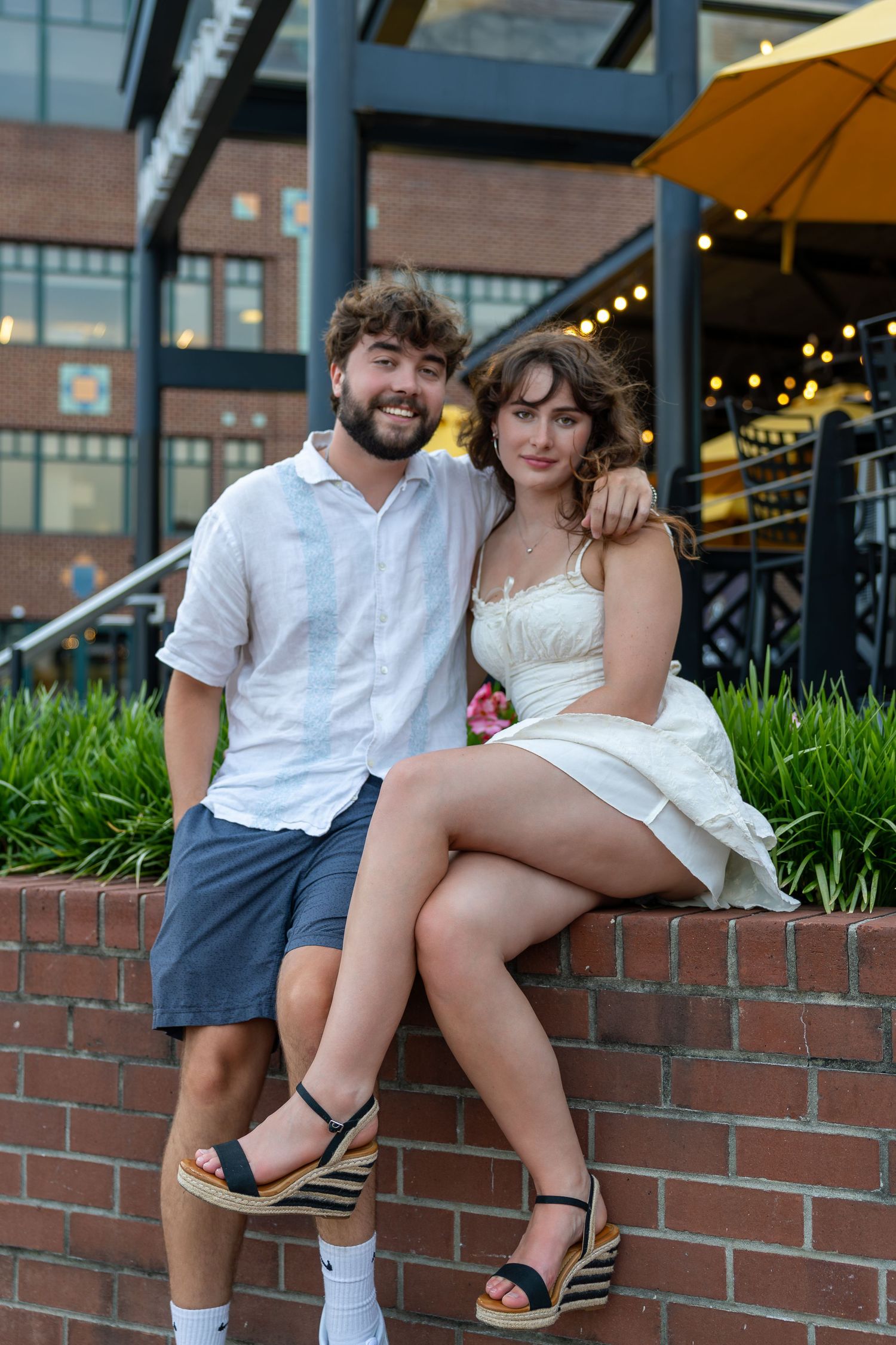 A smiling couple poses together on a brick wall outside a restaurant, woman in white dress with wedge heels, man in white shirt.