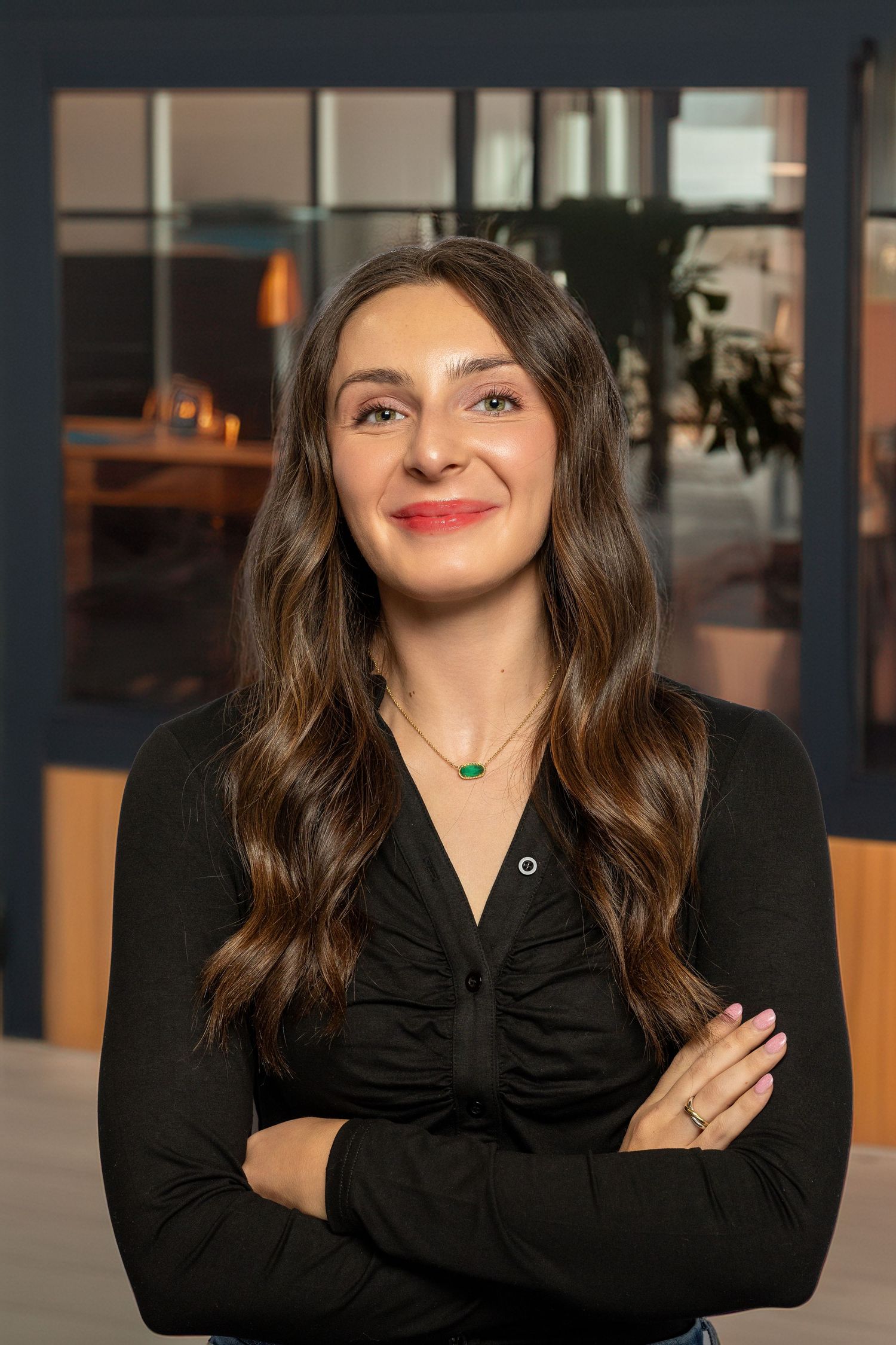 Professional woman with long brown hair in black top, arms crossed, smiling confidently in modern office setting.