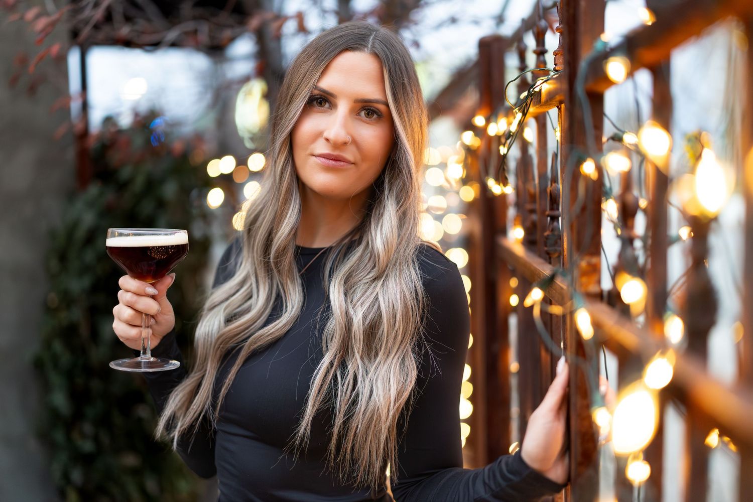 Woman in black sweater holding wine glass against blurred bokeh lights and greenery background.