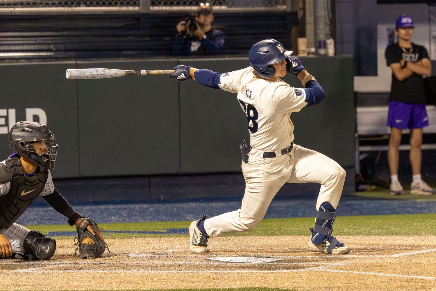 Baseball batter in blue uniform swings at pitch during night game as catcher crouches behind home plate.
