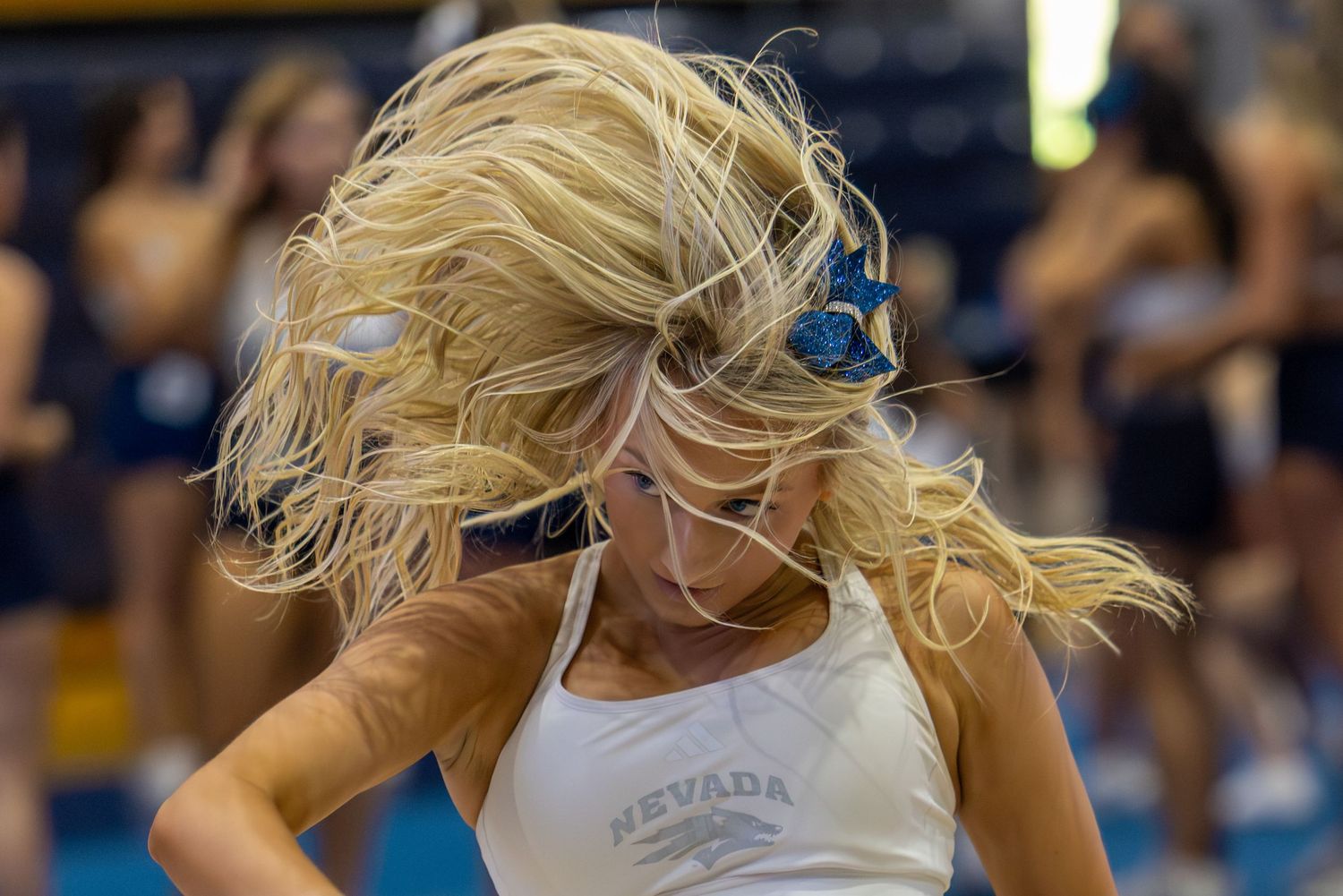 Nevada cheerleader with flowing blonde hair mid-dance routine in white tank top at indoor sporting event.