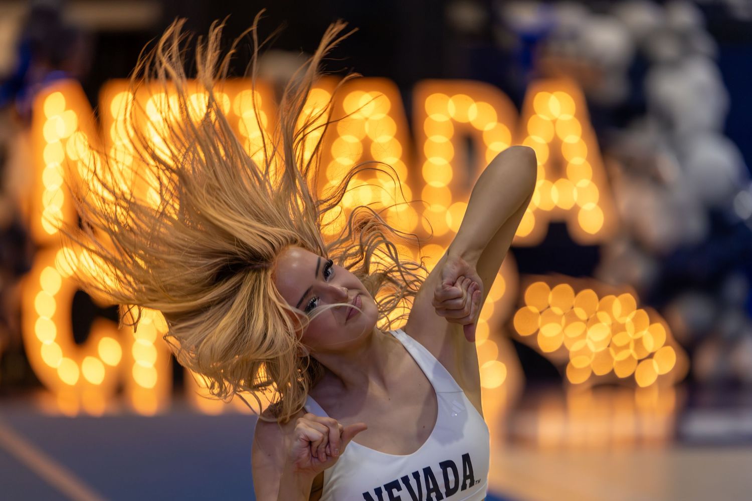 A blonde cheerleader in a Nevada uniform energetically performs a dance routine with illuminated Nevada letters in the background.