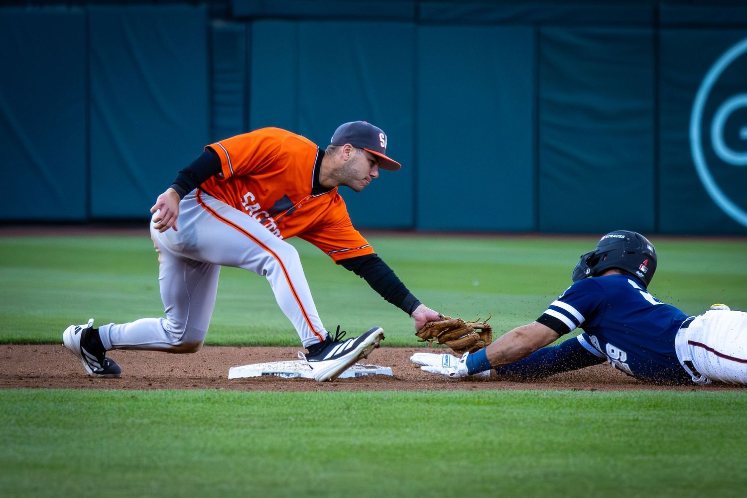 Baseball infielder in orange uniform tags out sliding runner in blue at second base during a game.