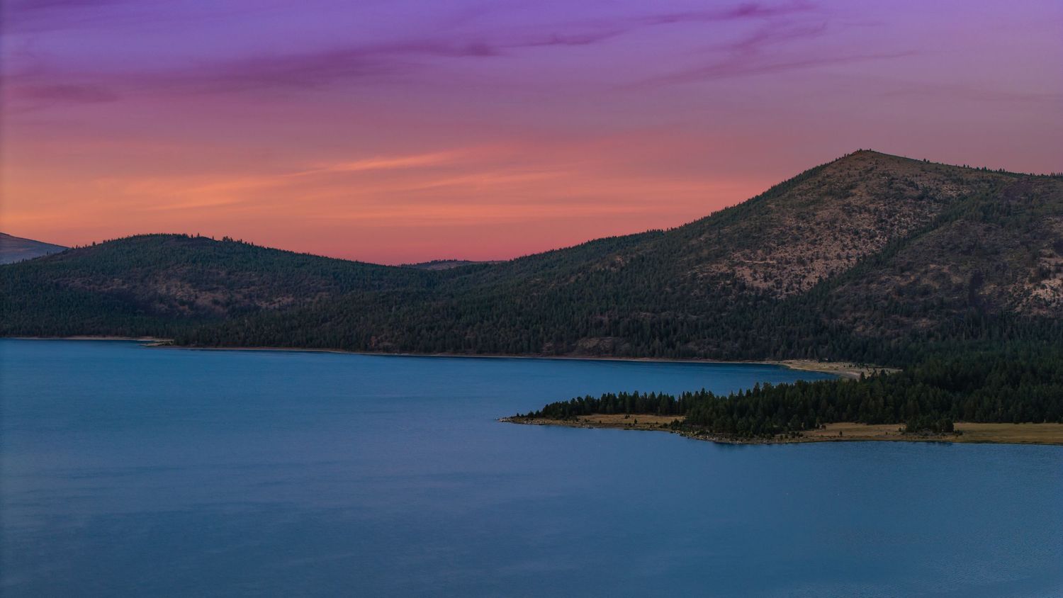 Panoramic view of a purple and orange sunset over a mountain lake with silhouetted hills in the background.