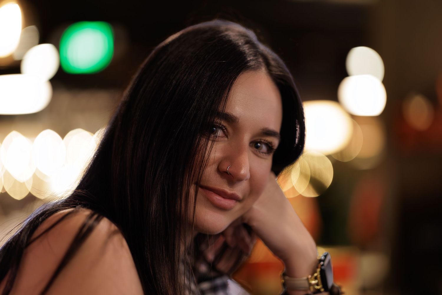 A woman in a dark sweater poses against a blurred background with bokeh lights.