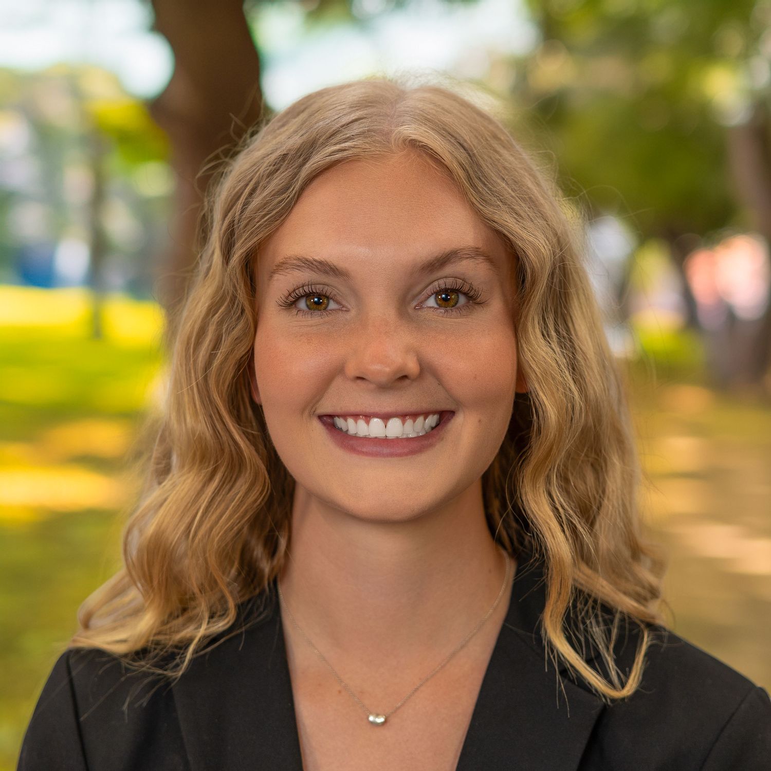 Young professional woman with blonde wavy hair smiling outdoors in a park setting, wearing a black blazer.