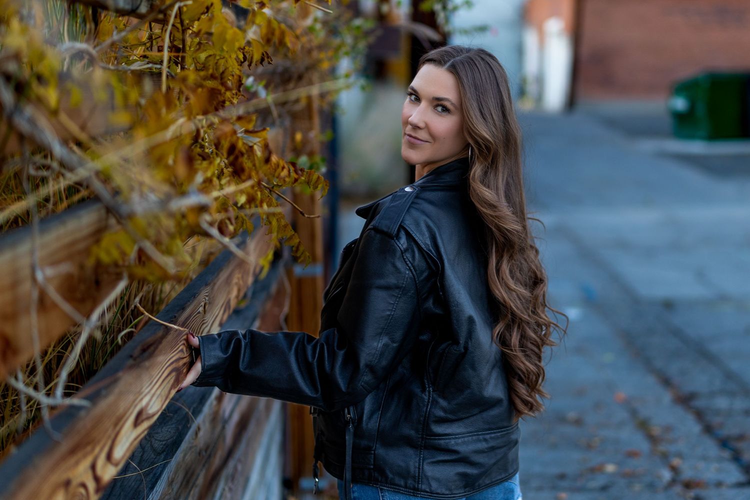 Young woman with long brown hair in a black leather jacket smiling over her shoulder on a brick sidewalk.