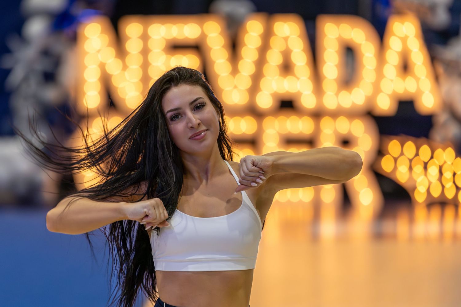 A young woman dances energetically in front of a glowing Nevada sign, wearing a white crop top.