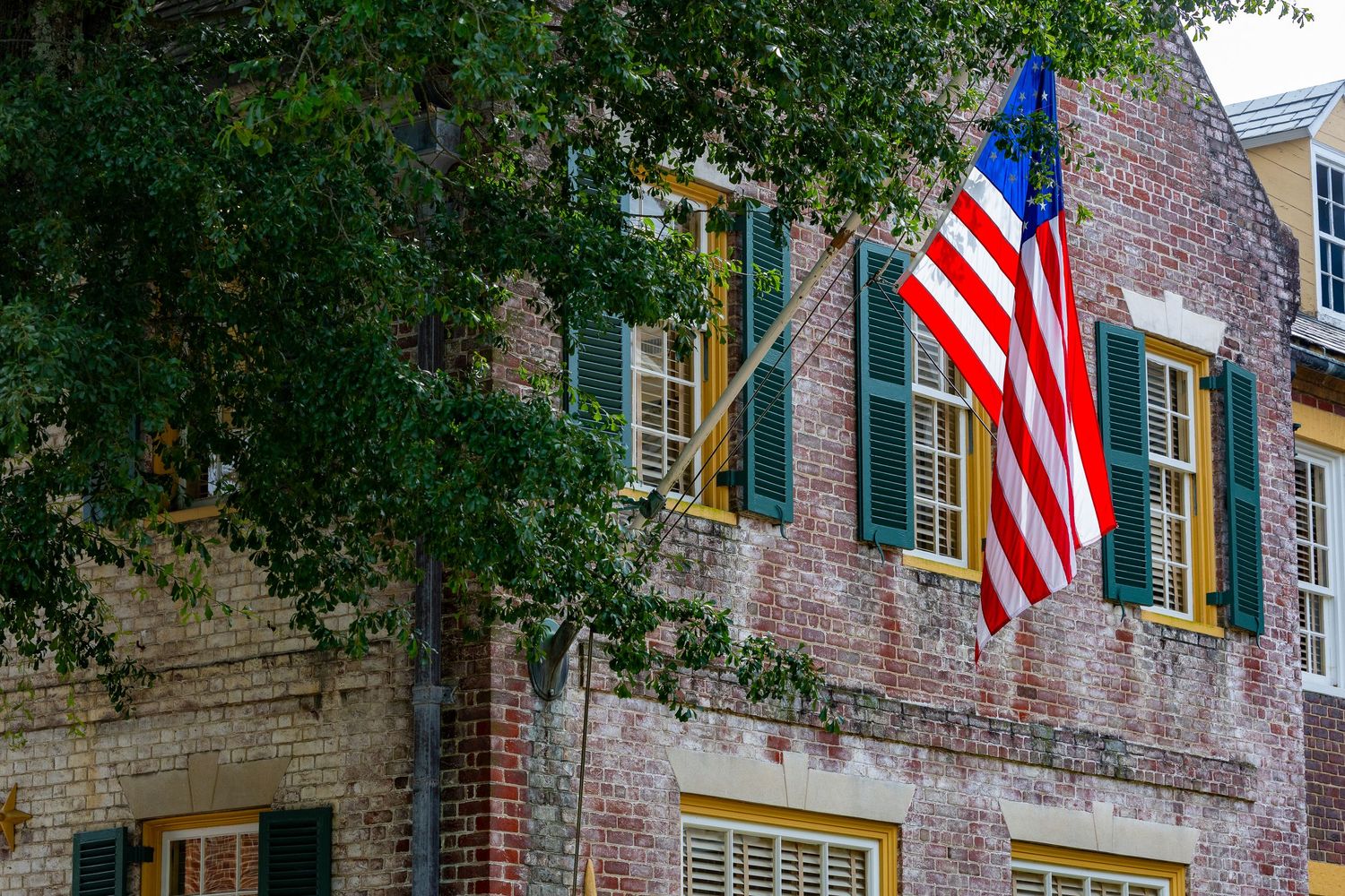 American flag displayed on a historic brick building with green shutters and yellow windows surrounded by trees.