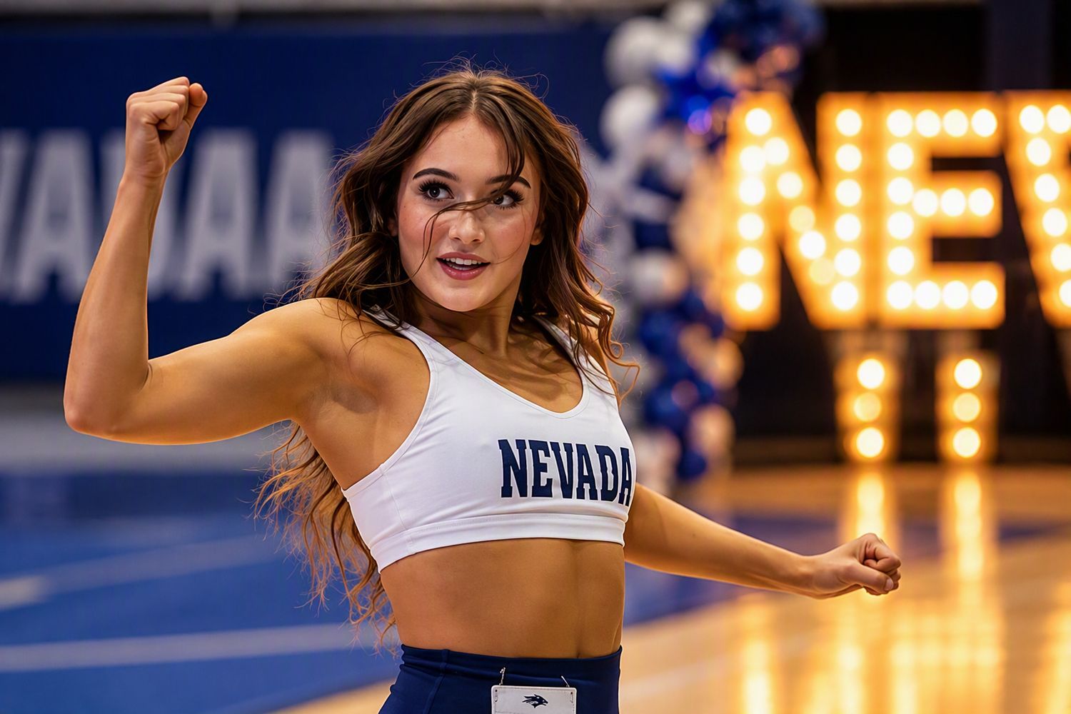 Nevada spirit squad dancer in a white crop top performs a pose with raised fist inside an arena with blue decorations and glowing lights.