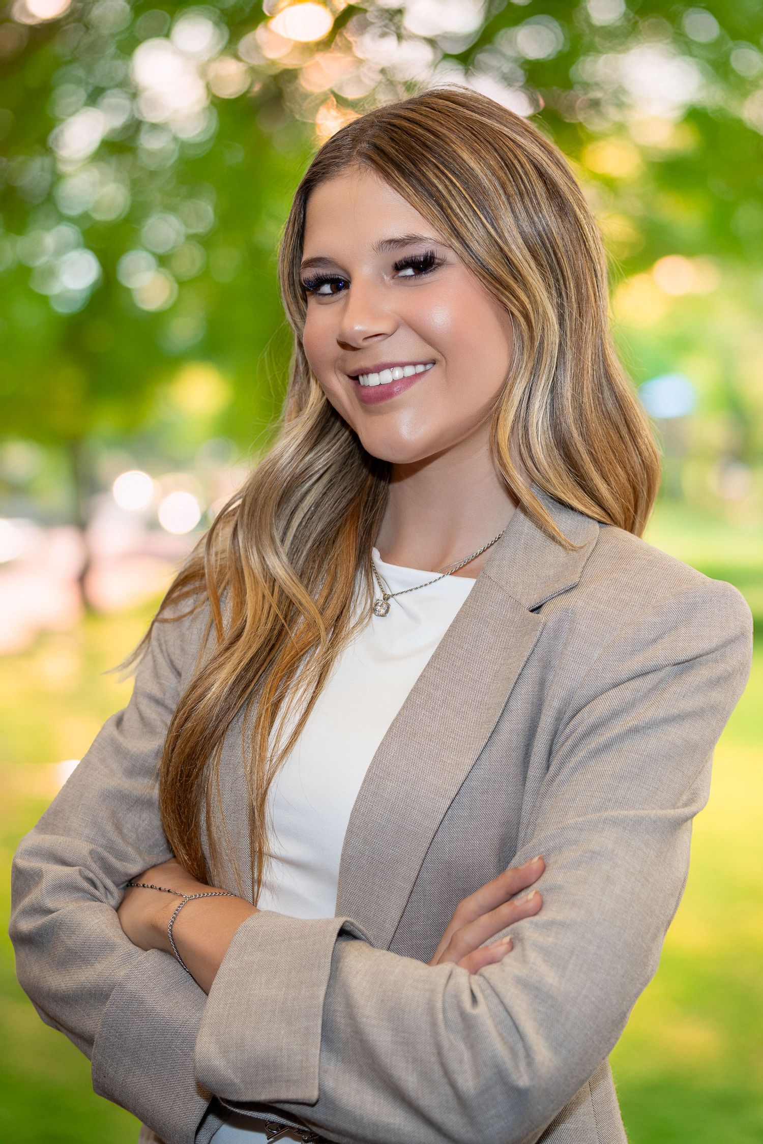 Young professional woman in beige blazer smiling confidently with arms crossed in a green outdoor setting.