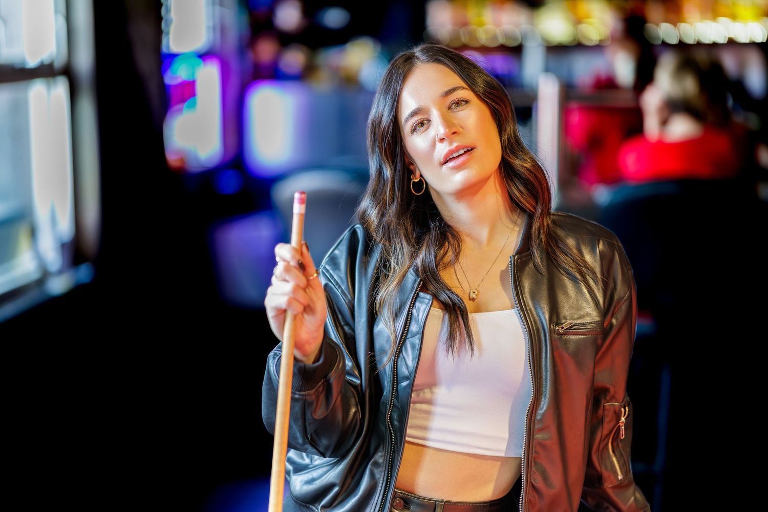 A person in a brown leather jacket and white crop top poses against colorful nightclub lighting.
