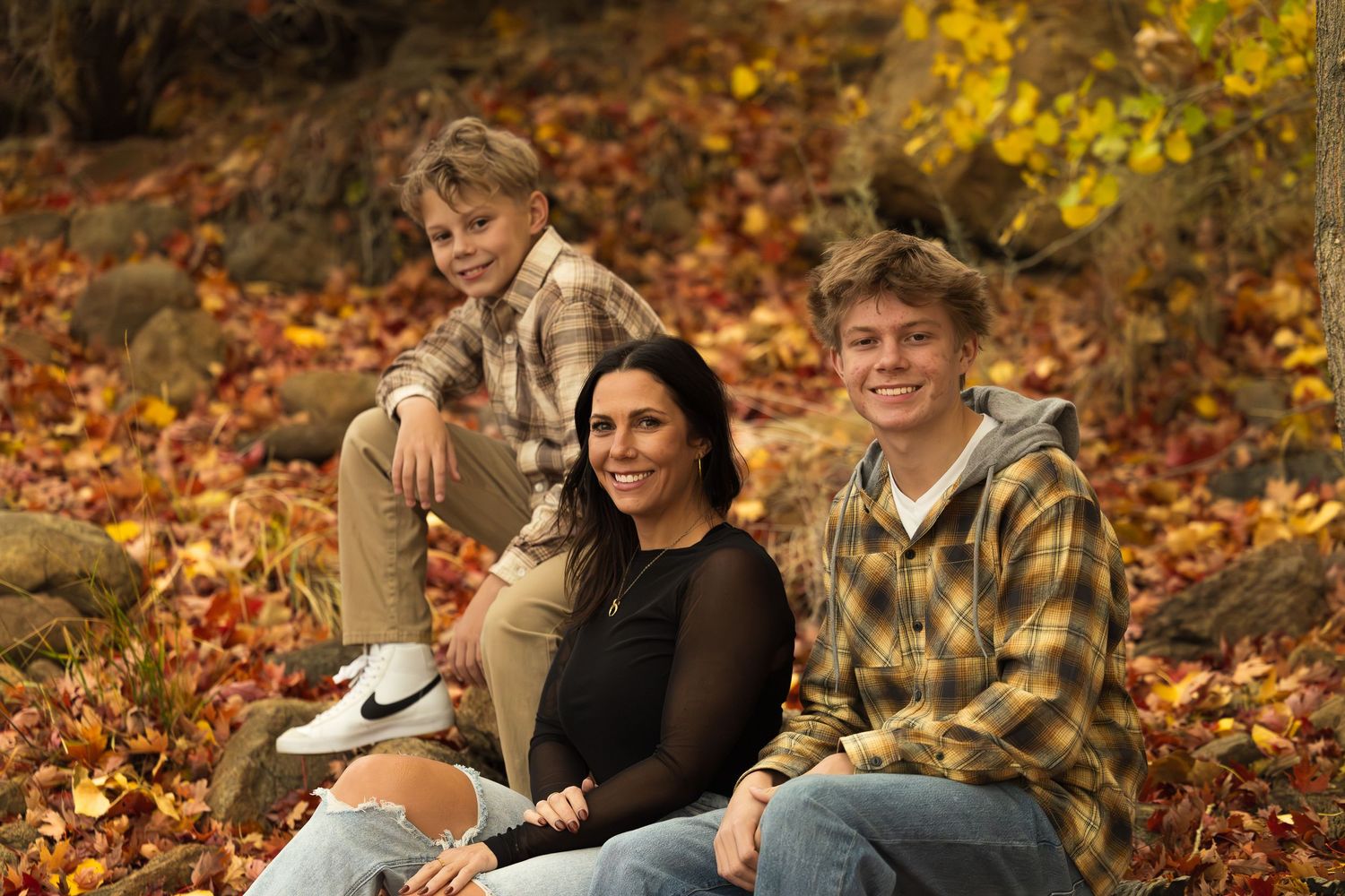 A group sits together on fallen leaves during an autumn photo session in a wooded area.