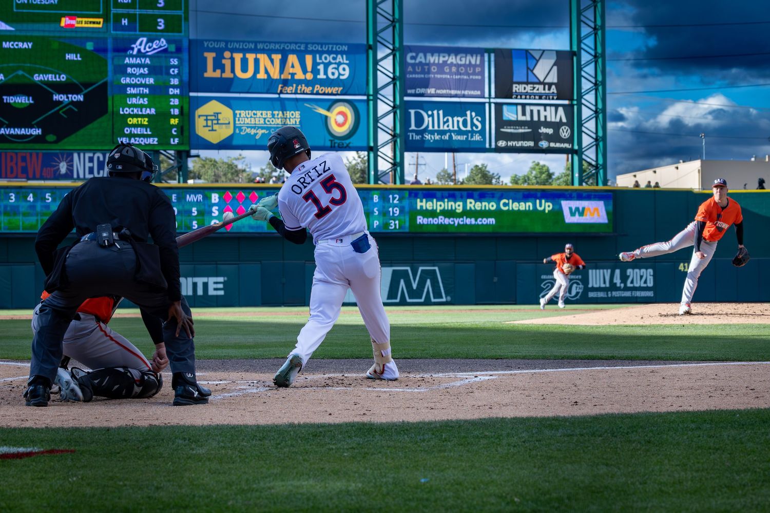 Baseball batter swings at pitch during minor league game with scoreboard and advertisements visible in background.