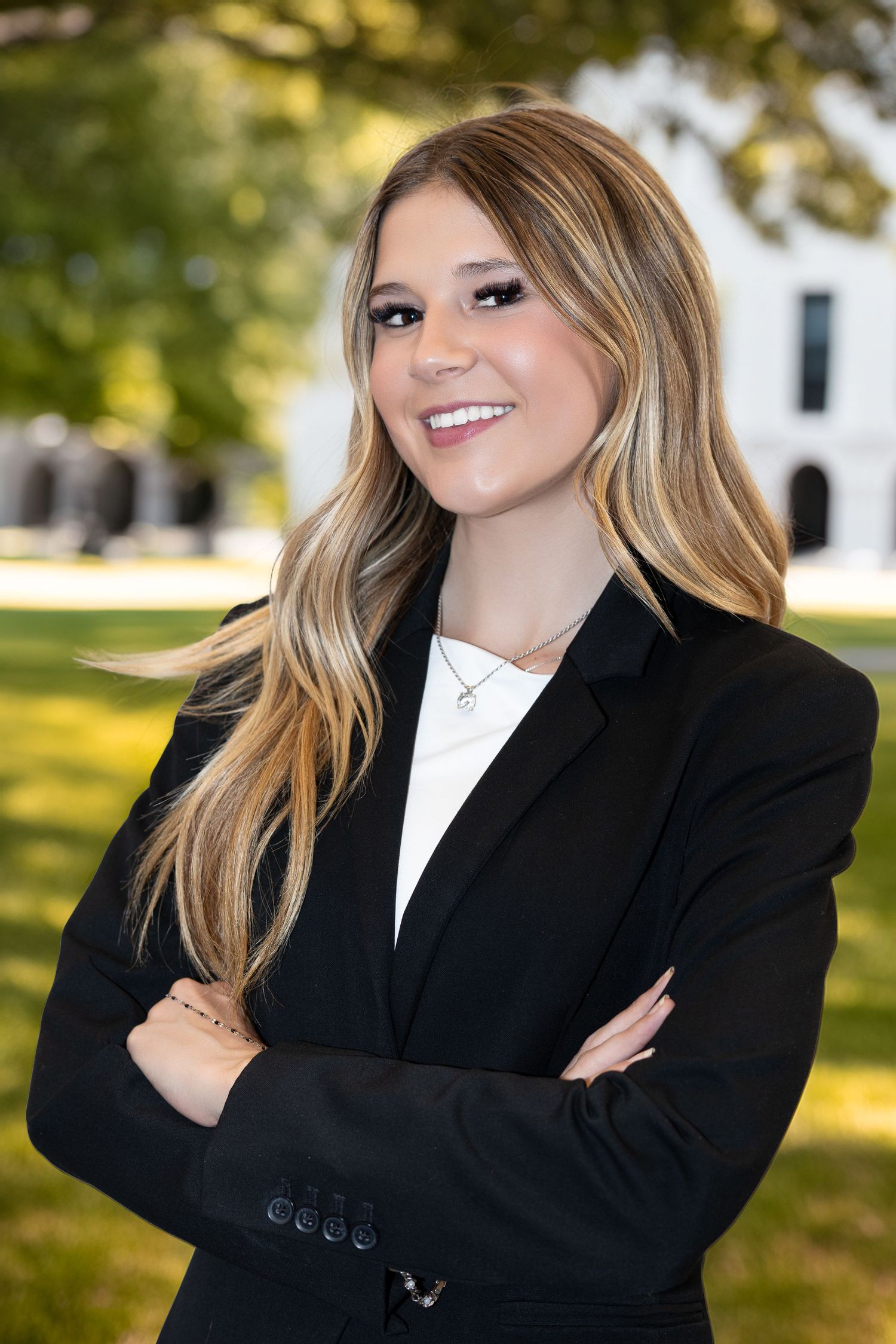 Young professional woman in black blazer smiling outdoors with arms crossed on a sunny campus.