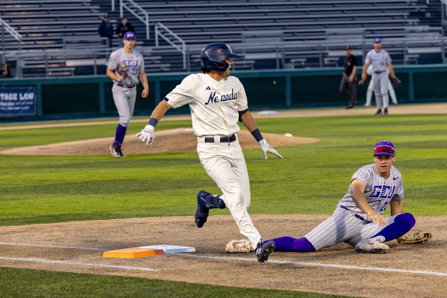 Baseball player runs to first base as fielder slides to make a play during a night game.