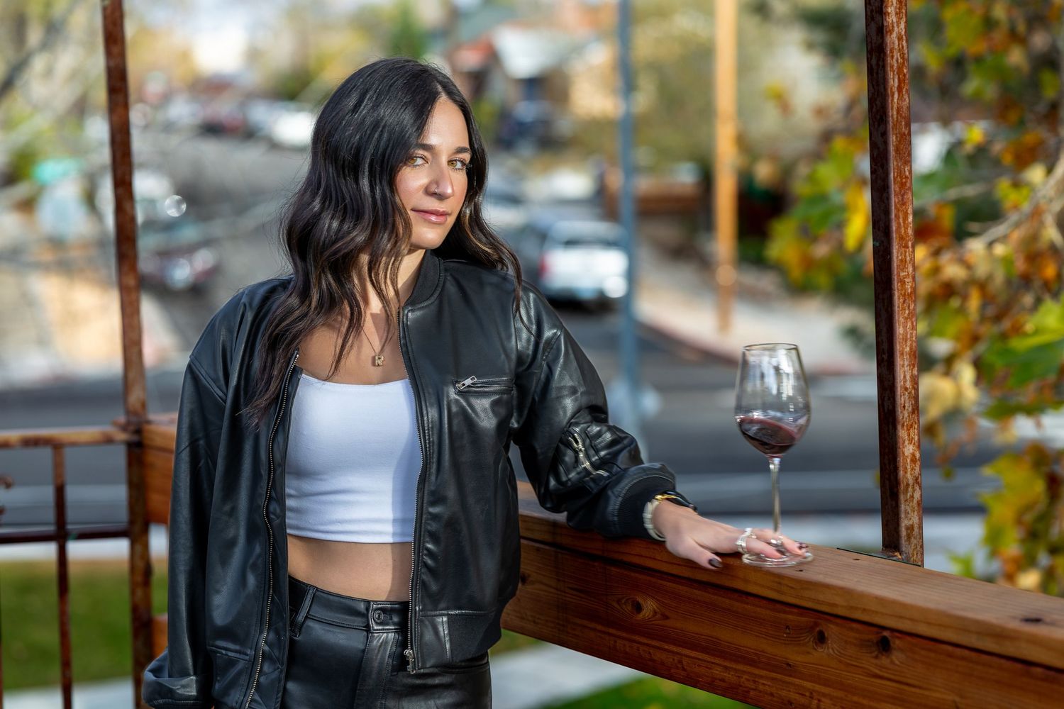A person in a black leather jacket sits at an outdoor table with a glass of red wine on an autumn day.