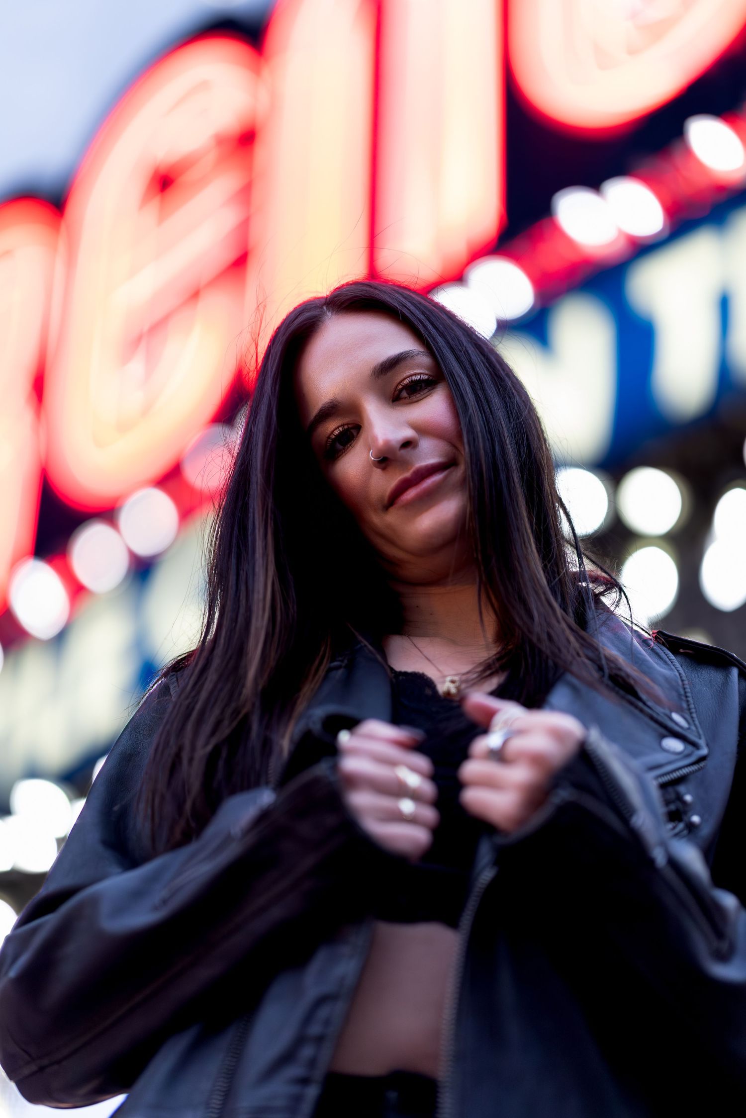 A person in a black leather jacket poses against blurred neon lights in an urban nighttime setting.
